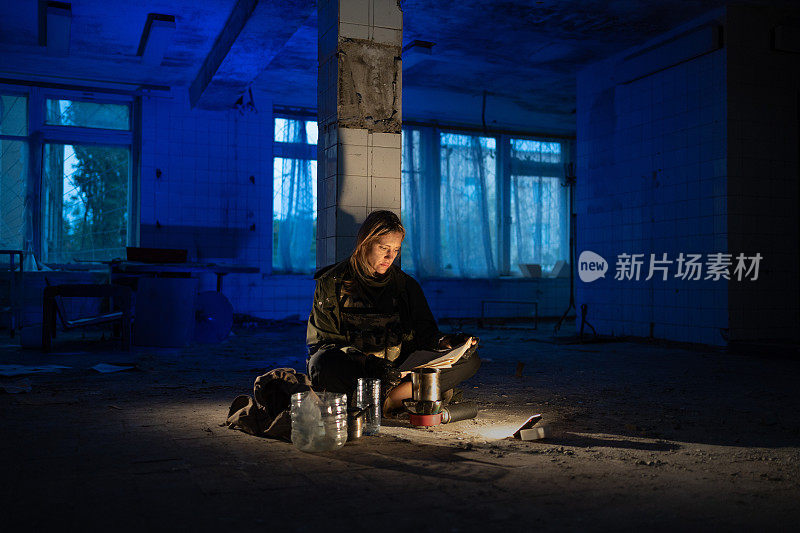 一个肮脏疲倦的女人晚上坐在一间废弃房间的地板上，拿着灯笼看报纸。出奇地。hiking in a post-apocalyptic world.